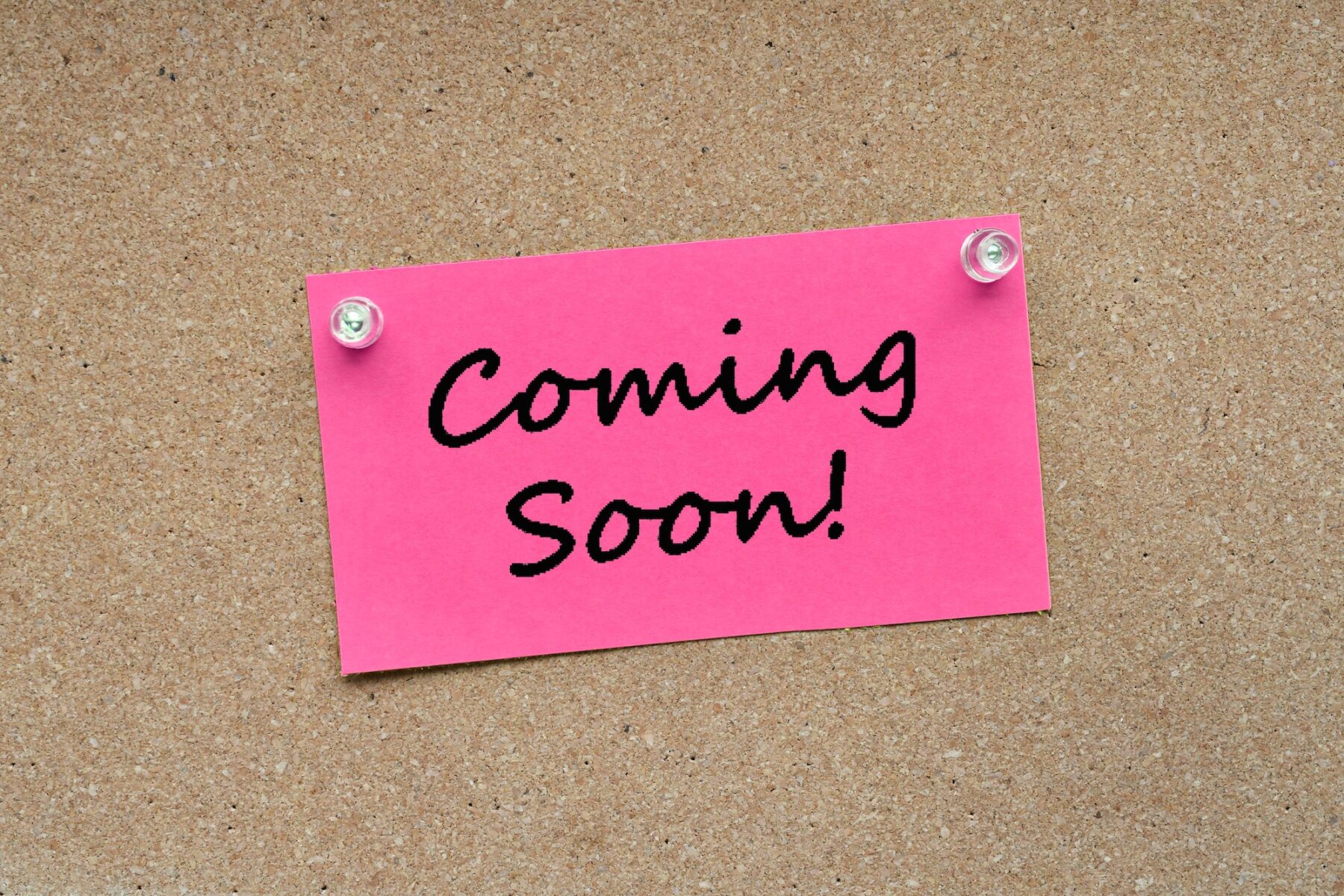 Pink note card pinned with push pins on a corkboard bulletin board with the words Coming Soon!